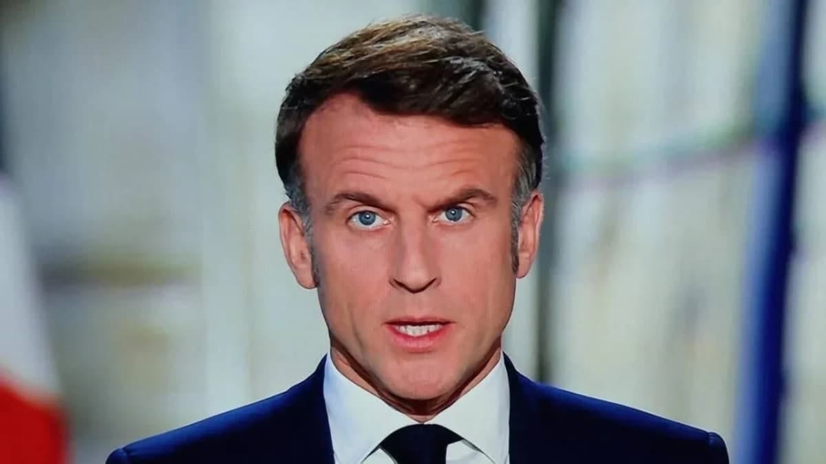 A close-up portrait of French President Emmanuel Macron wearing a dark suit, white shirt, and dark tie, speaking with a serious expression.