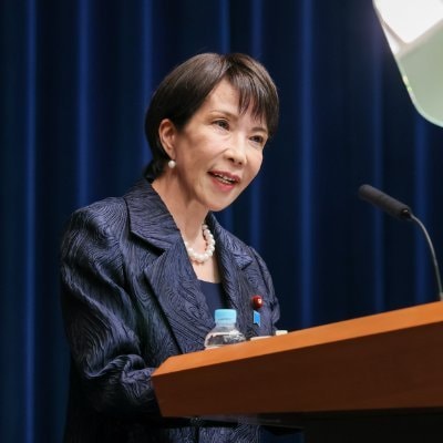 Here is everything for the photo from her X account:  *Title:* Sanae Takaichi Speaks Japanese Prime Minister Sanae Takaichi standing at a wooden podium speaking during an official press conference, wearing a navy-blue blazer and pearl necklace, with a water bottle in front of her.  